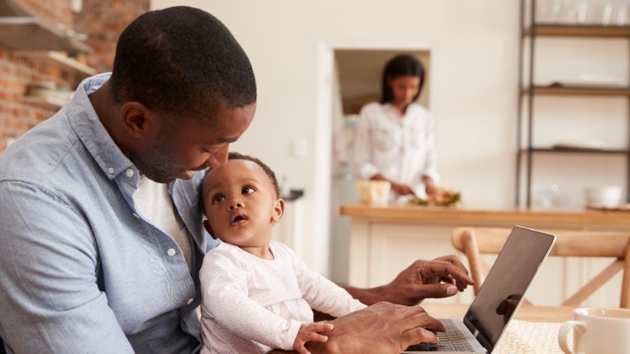 Parent with their baby at computer while person does laundry in the background