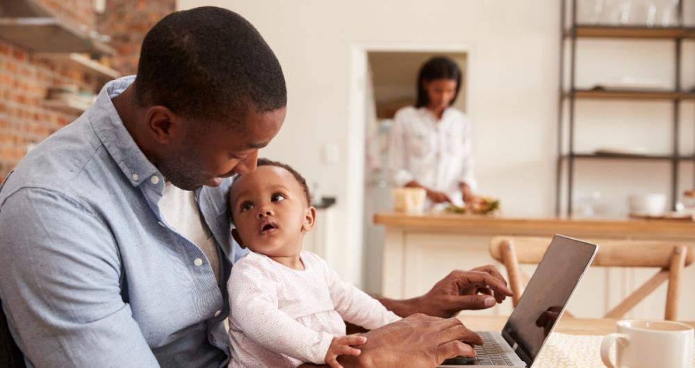 Parent with their baby at computer while person does laundry in the background